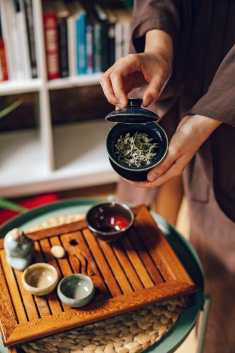 man holding a gaiwan with green tea leaves and bowls with tea pet on wooden board